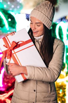 Winter concept. Happy young woman in hat holding christmas present box while standing on street.