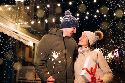 Two people celebrate New Year on the street. Close up portrait of young happy couple standing on the street hugging holding sparklers. Young couple in love holds sparklers in their hands.