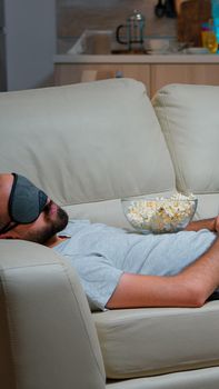 Man eye sleep mask falling asleep while watching movie, sitting in front of television. Caucasian male with beard looking entertainment series late at night in kitchen