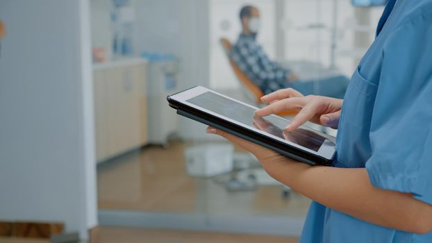 Close up of modern tablet with touchscreen and nurse hand using digital technology for development. Assistant holding device while stomatologist in cabinet consulting patient for oral care