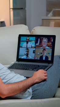 Tired man in pajamas sitting on sofa falling asleep while having online business videocall with teammates using laptop computer. Caucasian male on conference internet web communication