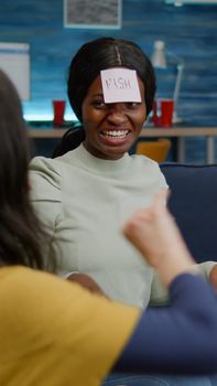 Multi-ethnic friends enjoying time together while relaxing on couch playing guess who game using sticky notes attaching on forehead. Mixed race people having fun in living room late at night
