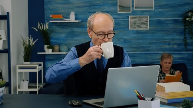 Tired elderly man sitting at desk and taking a sip of coffee, looking at laptop working from home workspace while his wife is reading a book in background. Focused old entrepreneur checking graphics