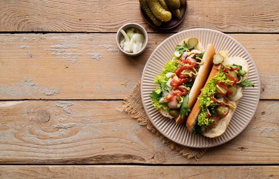 Hot dog with sausage, letuce, cucumber and onion on beige plate on wooden background. Top view. Flat lay.