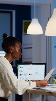 African businesswoman reading business reports from laptop in start up office late night typing new project. Focused employee doing overtime for job respecting deadline of project writing, searching