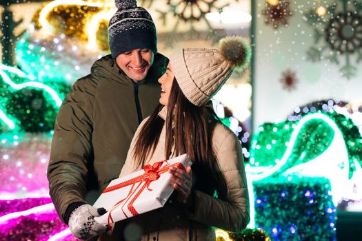 Close up of Young Beautiful Woman Hugging Man Holding Christmas Present. Pretty Girl Hugging With Boyfriend at Christmas Celebrating Holidays Together. Lovely Couple Stand. Gift Box Hugging Outdoors