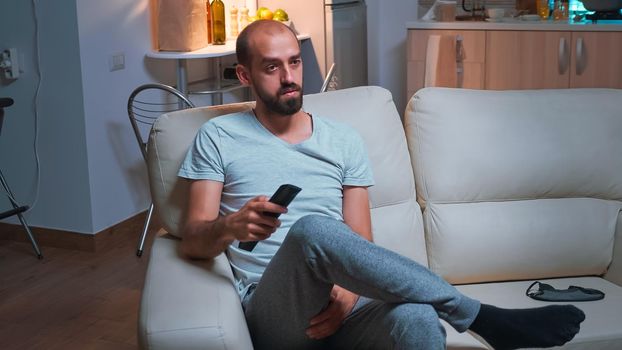 Portrait of concentrated man watching entertainment shows series sitting on couch in front of television. Caucasian male relaxing after long day at work late at night in kitchen