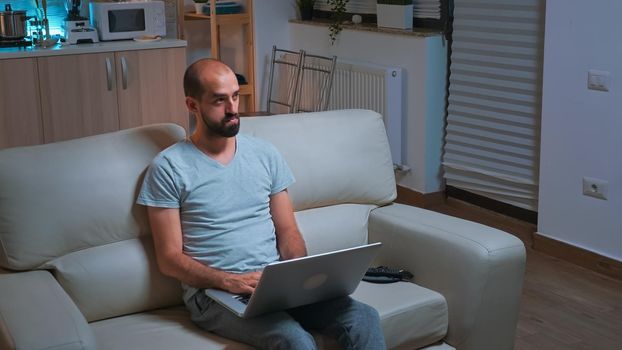 Caucasian male sitting in front of television while working at communication project before deadline on laptop. Tired man browsing on internet using network technology late at night in kitchen