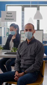 Portrait of businessman sitting at office desk wearing medical face mask against infection with coronavirus during global pandemic. Teamworkers working at business project maintain social distancing