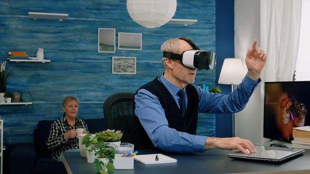 Senior man using virtual reality goggles in living room gesturing sitting in home workplace. Retired man experiencing vr headset while woman watching tv sitting on sofa in background.