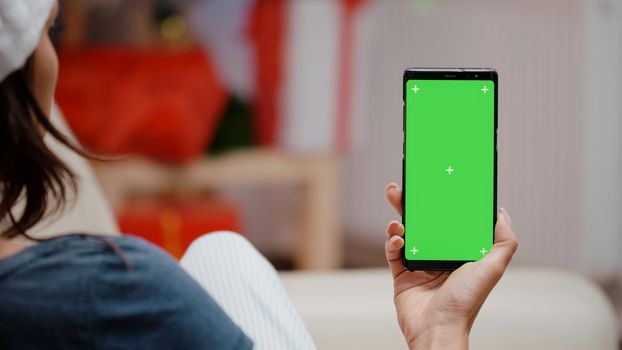 Close up of woman vertically holding green screen on smartphone in christmas decorated kitchen. Person looking at device with chroma key, isolated background and mockup template.