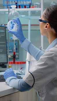 Innovation scientist studying petri dish with liquid bacteria while using modern computer with cell animation in laboratory. Woman with lab coat and glasses doing pharmaceutical research