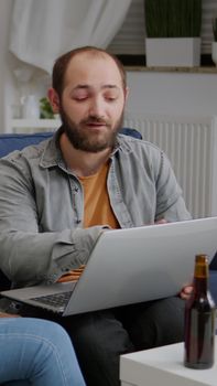 Multi-ethnic friends socializing while watching online funny videos on laptop resting on sofa. In background two womens drinking beer enjoying time spend together during entertainment party