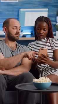 Interracial couple looking at smartphone screen at home. Young multi ethnic partners using modern device with technology while sitting on living room couch. Mixed race people with gadget