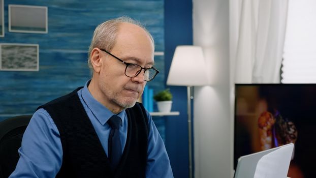 Close up of senior man writing at laptop working from home analysing financial project. Elegant retired employee with eyeglasses using modern technology reading typing on computer