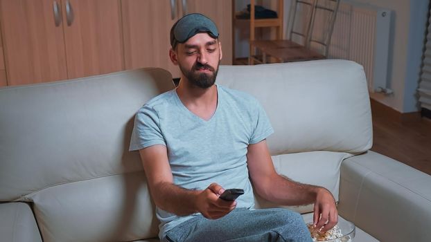 Interested man with beard relaxing in front of television while looking at entertainment movie using remote controle. Caucasian male lying on sofa eating popcorn late at night in kitchen