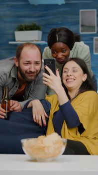 Mixed race friends chilling in living room during night party while watching online movie on phone display. Group of multiracial people laughing together, drinking beer relaxing on sofa