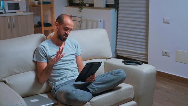 Focused man with beard sitting on couch in front of television while talking with teammates during online meeting videocall use tablet computer. Caucasian male using modern technology wireless