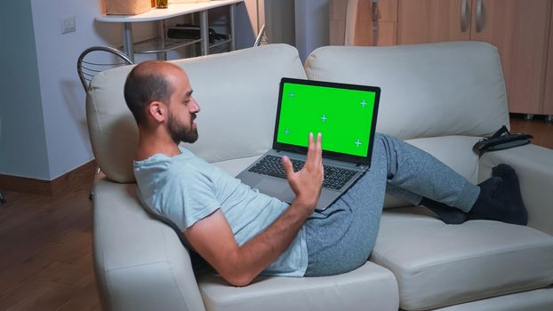 Tired man sitting on sofa while browsing marketing information using laptop computer with mock up green screen chroma key display. Caucasian male using modern technology wireless late at night
