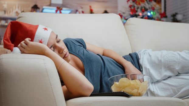 Close up of festive woman sleeping on sofa at television screen. Adult with santa hat resting on sofa while looking at movie on TV, feeling tired on christmas eve holiday. Sleepy person