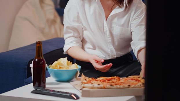 Young woman bringing pizza in living room on table to eat while watching television after work. Hungry person with snacks, fast food meal and bottle of beer enjoying takeaway food at home