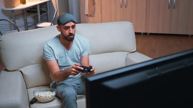 Upset pro gamer losing videogamer competition using wireless joystick during gaming competition. Disappointed player sitting in front of television on sofa making loser gesture late at night