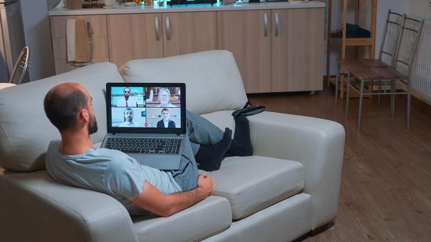 Caucasian male talking about business communication project during video call meeting use laptop computer. Focused man lying on sofa in pijamas using modern technology wireless late at night