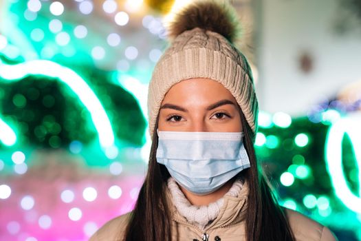 Close up portrait of Caucasian young beautiful female in medical mask standing in decorated xmas city. Crowd people sick covid-19. Epidemic coronavirus. Pandemic flu corona virus