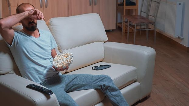 Concentrated man chilling on sofa in front of television watching movie series while eating popcorn. Caucasian male in pajamas looking at entertainment shows late at night in kitchen