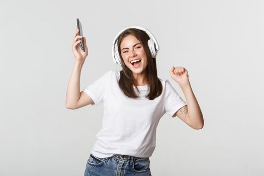 Happy beautiful brunette girl dancing and listening music in wireless headphones, holding smartphone.