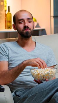 Man late at night in his apartment looking at TV entertainment, laughing and having fun while eating popcorn snack. Smiling caucasian person leisure time on the sofa in front of television
