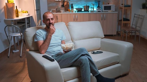 Man with beard watching tv show using remote control to change channels on television. Caucasian male relaxing on couch with popcorn bowl in hands looking entertainment series at late at night