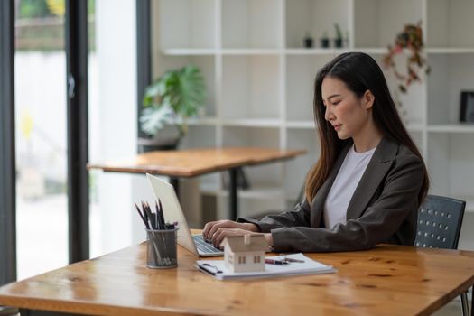 Real estate agent hand typing laptop keyboard with house model and keys, business asian woman working at home office