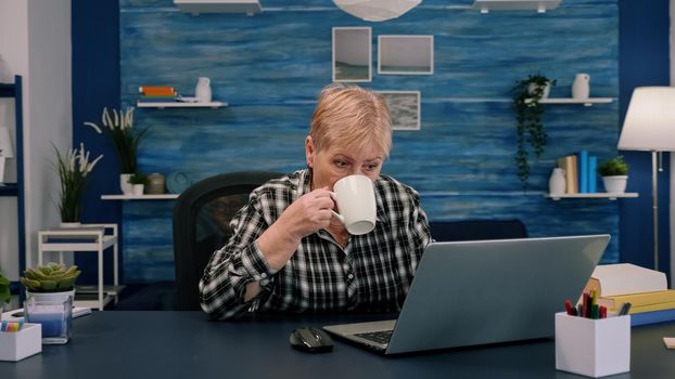 Senior woman enjoying a cup of coffee while working on laptop in living room while husband sitting on the couch and reading book in background. Mature older lady watching online business training