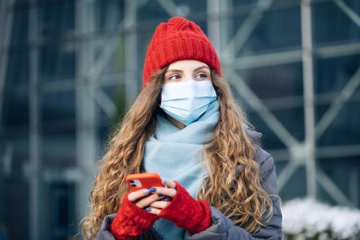 Joyful young woman texting on cellphone. Curly female standing on winter street. Caucasian beautiful curly female in mask browsing on smartphone while standing on street.