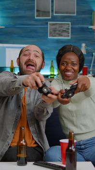 Two women losing online videogames using joystick playing for gaming competition. Multi-ethnic friends drinking beer, socialising, having fun together sitting on couch late at night