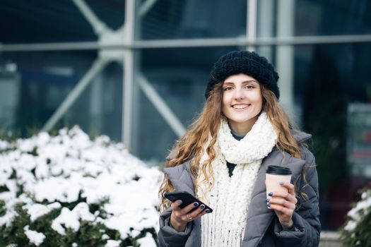 Curly female using smartphone standing outside. A smiling happy girl employee typing on a cellphone. Winter concept. Joyful young female in good mood typing and scrolling on smartphone outdoors.