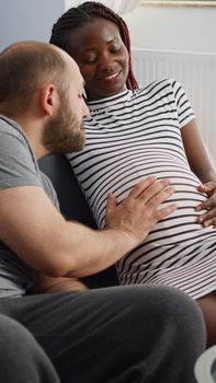 Married interracial couple bonding over child sitting at home. Young multi ethnic parents touching baby bump and talking about parenthood. Black pregnant woman and caucasian man