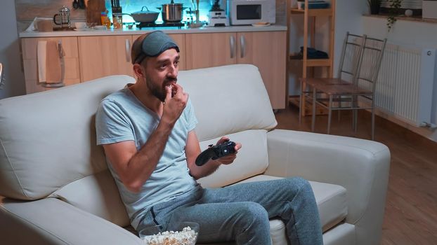 Frustrated man with sleep mask losing videogame competition using wireless controller. Disappointed pro gamer lying on sofa in front of television late at night in kitchen