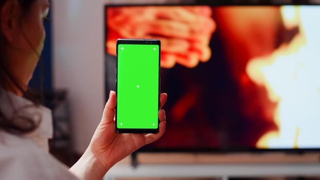 Close up of woman vertically holding green screen on smartphone at home. Person with isolated background and mockup template for chroma key while movie playing on TV in background