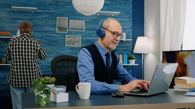 Senior old man listening music on headphones while working on laptop in living room and wife drinking coffee in background. Retired man using modern technology reading typing, searching on computer