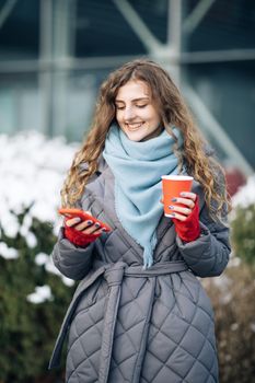 Female portrait. Walking look around on the street city. Being online, social networks. Modern stylish curly woman with cup coffee, uses her smartphone .