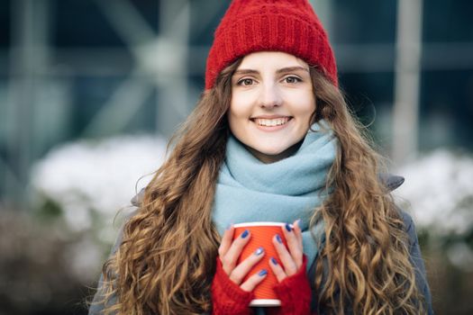 Portrait of nice-looking curly caucasian elegant young woman looking into camera. Female portraits. Girl in a red warm hat with a cup of coffee in her hands stands on the street in winter