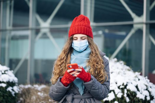 Portrait of Caucasian beautiful curly female in mask browsing on smartphone while standing on street. Joyful young woman texting on cellphone. Curly female standing on winter street