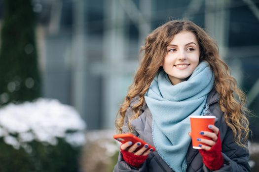 Portrait of happy beautiful curly woman with coffee cup takeaway use mobile phone texting messages. Walking look around on the street city.