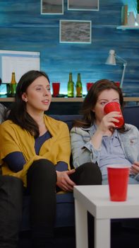 Multiracial friends sitting on couch watching fun movie on television during home party late at night in living room. Group of mixed race people drinking beer, eating snacks enjoying time together