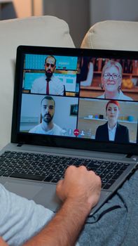 Over shoulder view of man in pajamas having online conversation with teammates working at online technology project using laptop computer, videoconference web internet communication