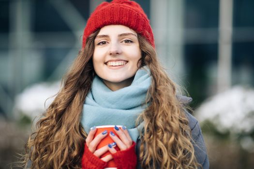 Cheerful gorgeous curly brunette in winter fashion holding coffee cup on urban background. Concept happiness, winter holidays, christmas, beverages and people. Curly female standing on winter street.