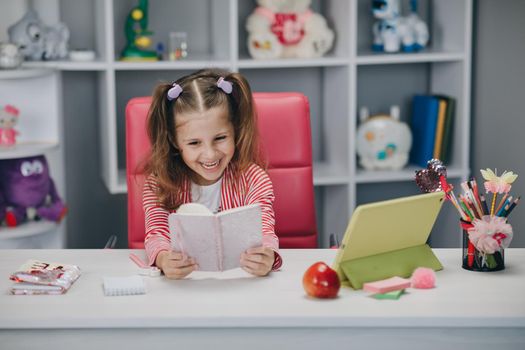 Self isolation and online schooling. Distance learning, school online. Portrait of a preschool girl looking at the book and smiling.