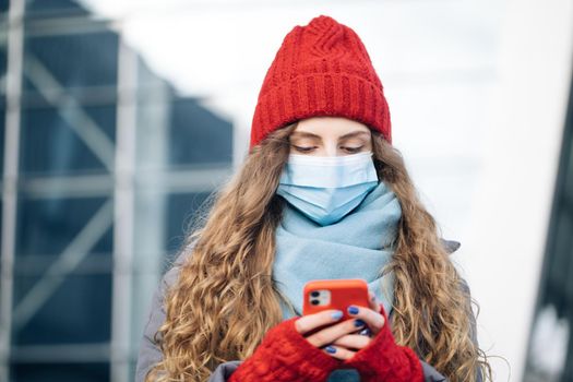 Curly female standing on winter street. Caucasian beautiful curly female in mask browsing on smartphone while standing on street. Joyful young woman texting on cellphone.